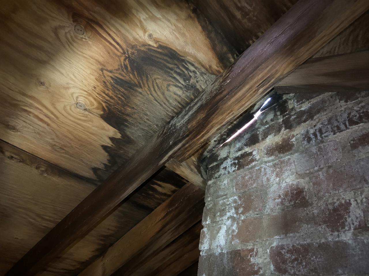 Water-stained roof decking and rafters inside an attic with daylight showing at the chimney