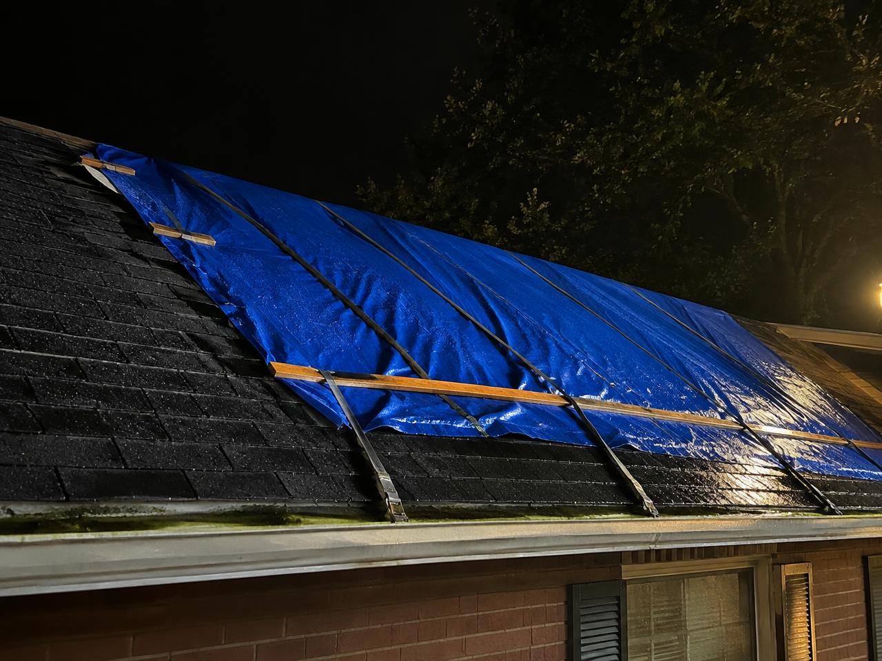 Blue emergency tarp strapped over a wet shingle roof at night under a work light